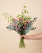 Eine Hand hält den Morgenlicht-Strauss mit rosa Ranunkeln, pfirsichfarbenen Rosen, Hagebutten und Eukalyptus vor einem warmen beigen Hintergrund.