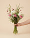 Eine Hand hält den Blumenstrauß 'Du bist so toll' in der Größe Classic mit weißen Rosen und rosa Nelken vor beigem Hintergrund.