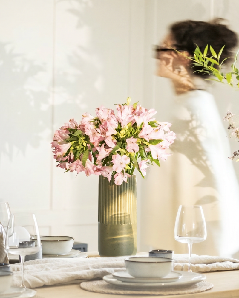 Dunkelgrüne feey Keramik-Vase 'Rilio' mit rosa Alstroemerien-Strauß auf einem gedeckten Esstisch, im Hintergrund eine Person