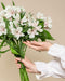 Eine Person in einer weißen, langärmeligen Bluse hält einen Strauß weißer Alstroemeria mit grünen Blättern, die vor einem einfarbig beigen Hintergrund fröhliche Stimmung verbreiten.