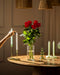 Eine Hand zündet eine weiße Kerze auf einem Holztisch an, mit Roten Rosen in einer Vase und leuchtenden grünen und weißen Kerzen unter warmen Hängelampen - eine wahre Liebeserklärung.
